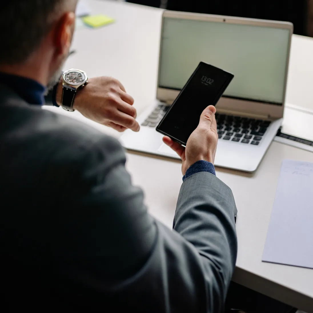 Man achter een laptop kijkt op zijn horloge terwijl hij zijn smartphone vasthoudt